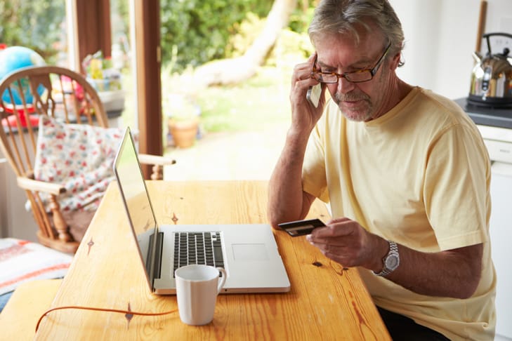 man with beard and glasses in yellow t-shirt sitting at table with computer and credit card out, on the phone.