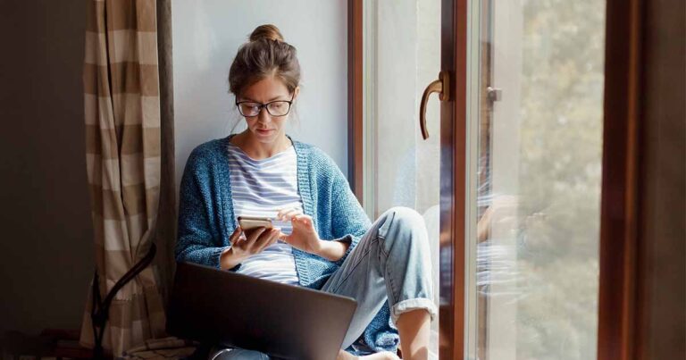 woman sitting near window with smartphone and laptop