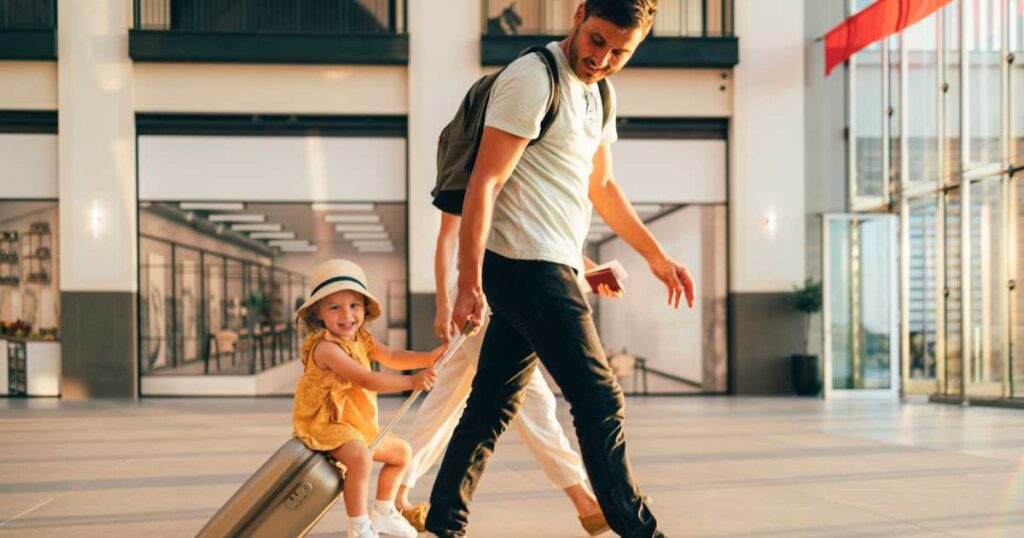 Cheerful husband and his anonymous wife walking with their little girl sitting on luggage at the airport.