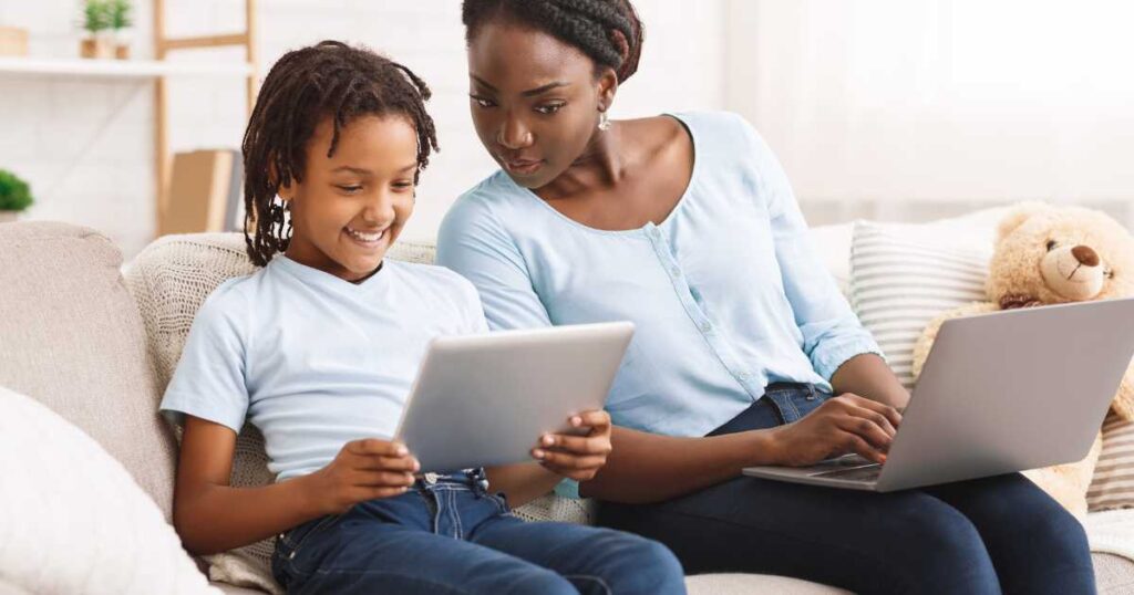 Black mother watching her daughter's activity online. Sitting together on a couch with a laptop and a tablet.